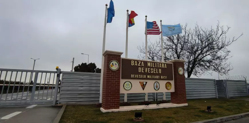 Imaginea arată intrarea în Baza Militară Deveselu, cu un panou mare pe care este inscripționat numele obiectivului în română și engleză. Deasupra sunt arborate mai multe steaguri, inclusiv cel al României și al Statelor Unite, iar zona este împrejmuită cu gard metalic de securitate.