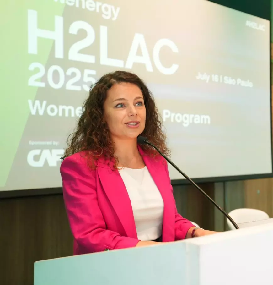 O femeie în fața unui pupitru cu microfon, îmbrăcată în bluză albă și sacou roz