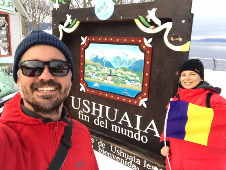 Un bărbat și o femeie, cu geci roșii și căciuli negre în cap, pozează cu un semn pe care scrie USHUAIA. Femeia tine in mana un steag al României
