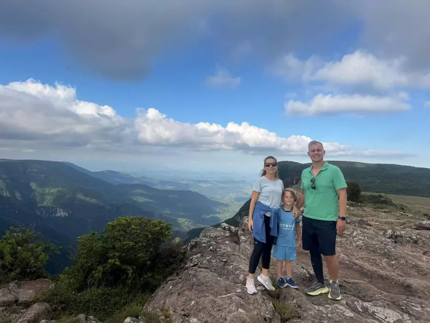 O familie formata din mama, tata si copil, imbracati in haine sport, poză dintr-o vacanță. In spate se vede un peisaj montan