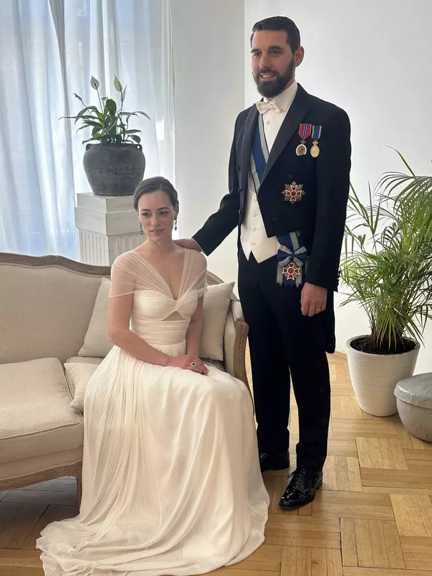 Un cuplu, el in frac negru, camasa alba si pantaloni negri, sta in picioare langa o canapea, ea, imbracata intr-o rochie alba, sta pe canapea