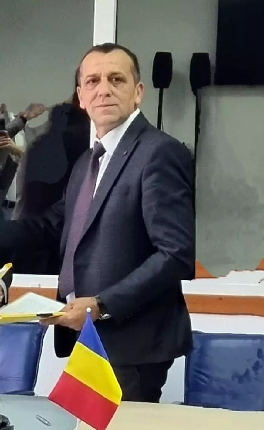 Un barbat imbracat in costum albastru inchis este intr-un birou langa o masa pe care este steagul Romaniei