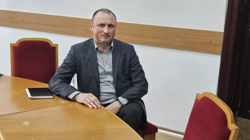 Un barbat in costum gri sta la o masa si acordă un interviu