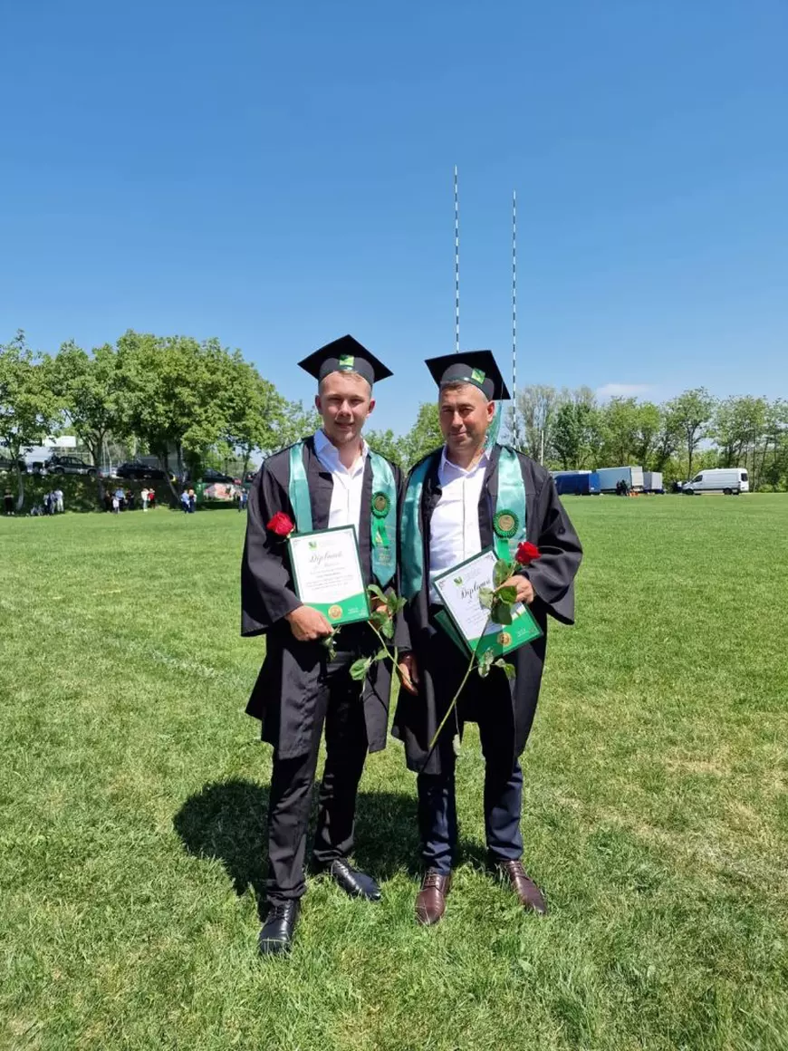 Doi bărbați pe un teren înverzit, la absolvirea facultătii, cu tocă și robă negre și eșarfe verzi, tin cate o diplomă și cate un trandafir roșu în mână