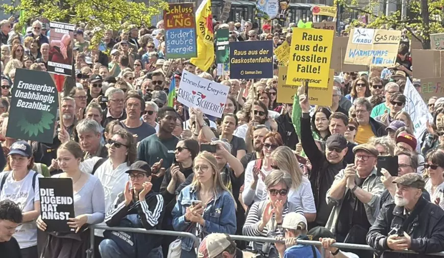 Imaginea surprinde o mulțime densă de oameni care ocupă o stradă urbană mărginită de clădiri multietajate cu ferestre înalte și copaci subțiri, totul fiind redat într-o perspectivă de sus care se extinde spre fundal. Participanții sunt îmbrăcați în haine de zi de diverse culori și poartă numeroase pancarte albe sau colorate cu mesaje scrise și desene, alături de steaguri galbene, albastre și verzi care flutură deasupra capetelor.