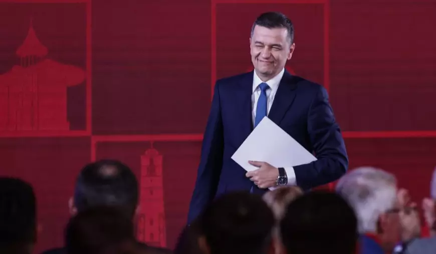Sorin Grindeanu, îmbrăcat în costum, cămașă albă și cravată albastră, în timp ce face cu ochiul și ține într-o mână o foaie albă de hârtie