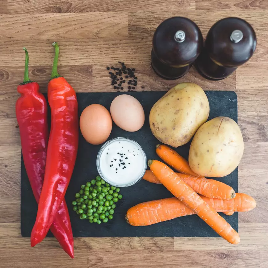 Ingrediente alimentare pe o tablă neagră. Sare și piper, cartofi, morcovi, sos alb, mazăre, ardei și ouă.