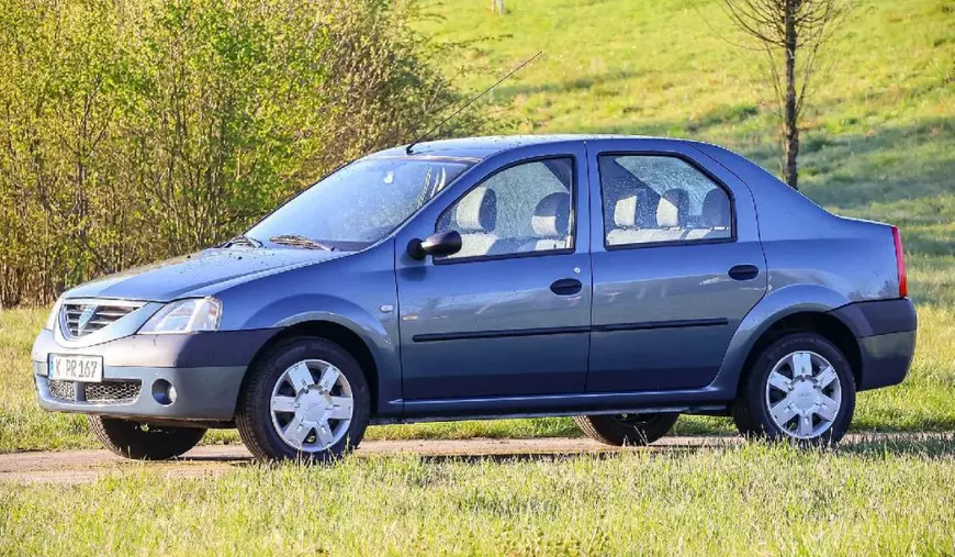 Imaginea prezintă un autoturism Dacia Logan albastru din prima generație, parcat pe marginea unui drum, cu iarbă și arbori în fundal.