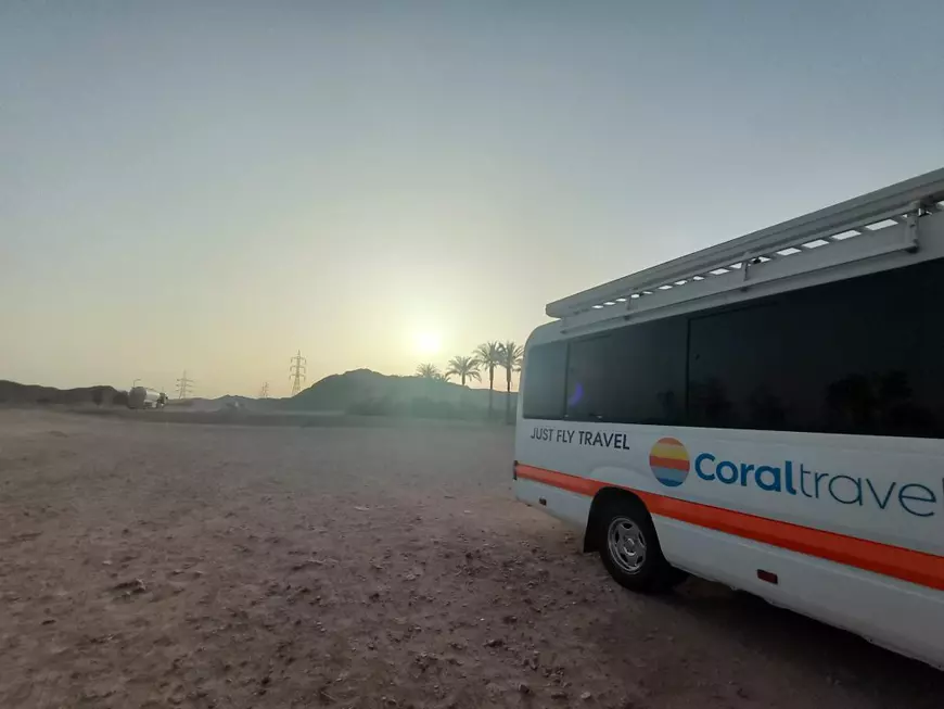 vedere a dunelor de nisip din Luxor, la răsărit, cu palmieri în depărtare. În dreapta imaginii este un autobuz alb, de transport pasageri, cu logo-ul Coral Travel și textul „Just Fly Travel”