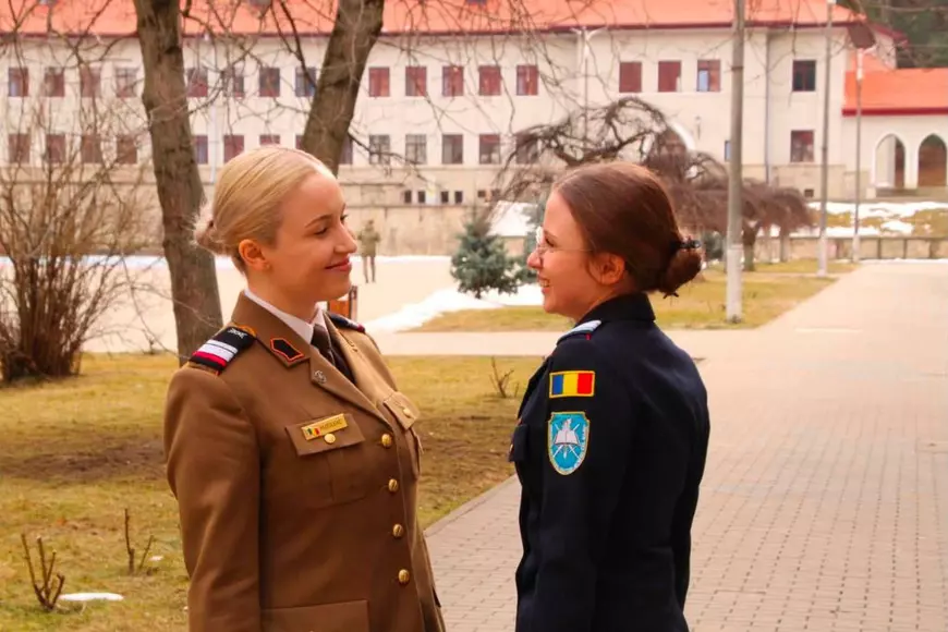 Două fete, îmbrăcate în haine militare se uită una la cealalta, in curtea unei instituții